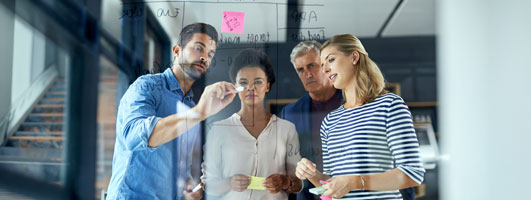 Cuatro personas escribiendo y observando notas adhesivas sobre una superficie de cristal